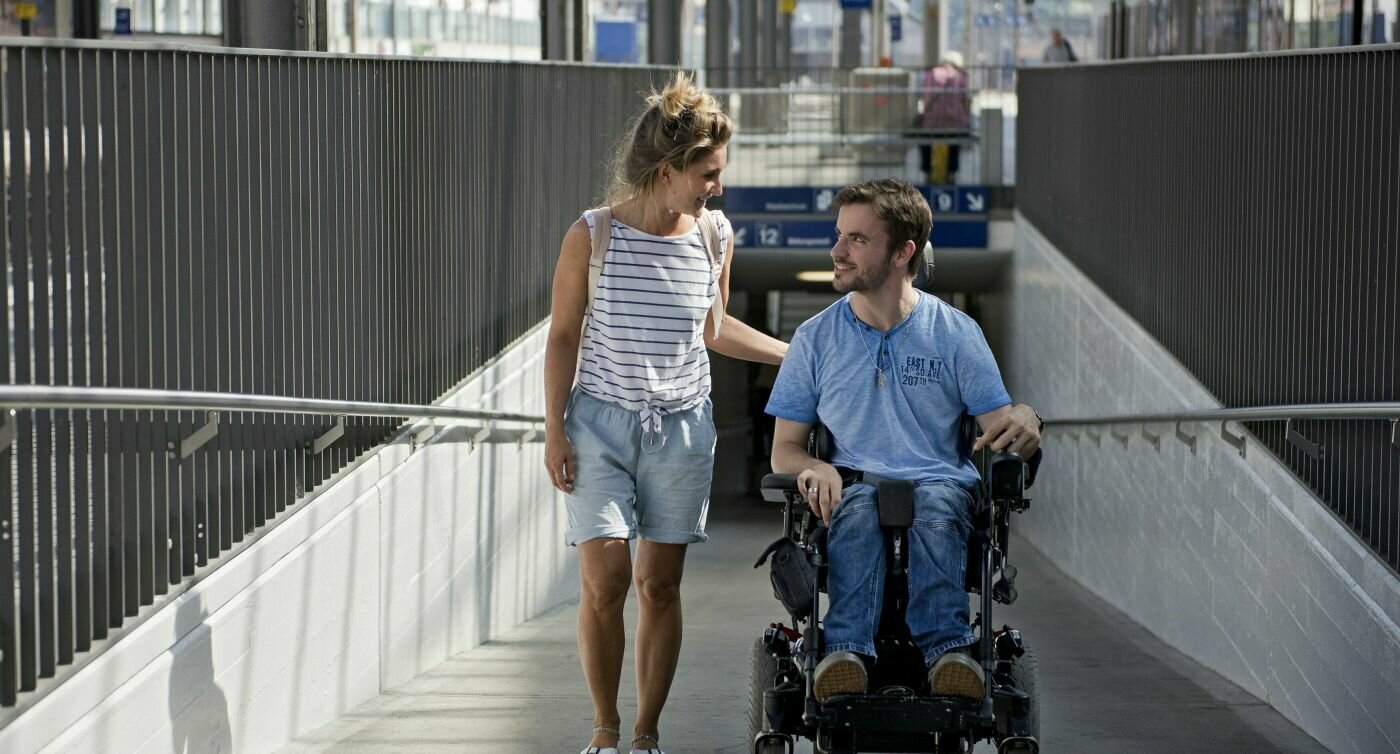 Uomo in sedia a rotelle accompagnato da una donna su una rampa in stazione