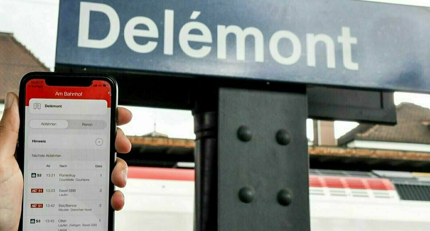 Cartello della stazione di Delémont con Beacon, davanti al quale qualcuno tiene in mano il suo cellulare