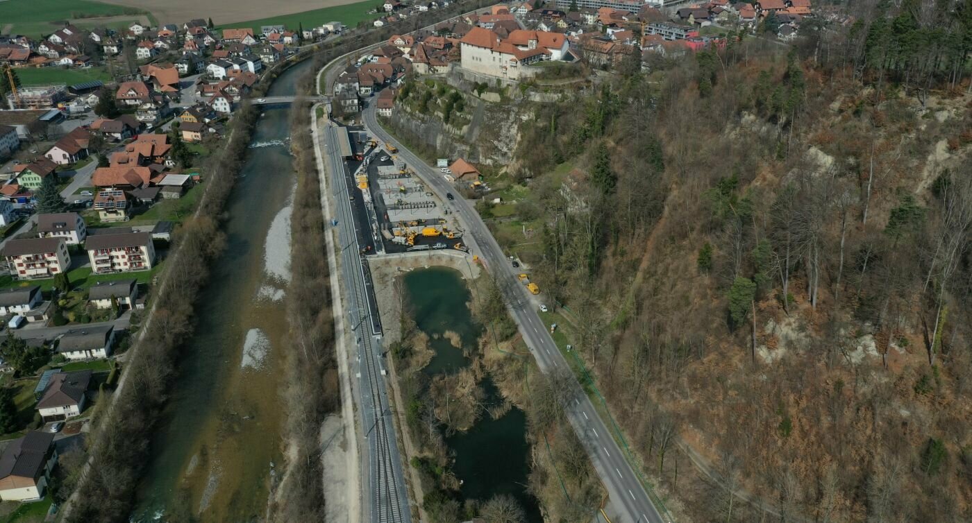 Luftaufnahme des Bahnhofs Laupen. Zu sehen ist ein Fluss, die Gleise und die Hauptstrasse umgeben von Wald und dem Dorf.