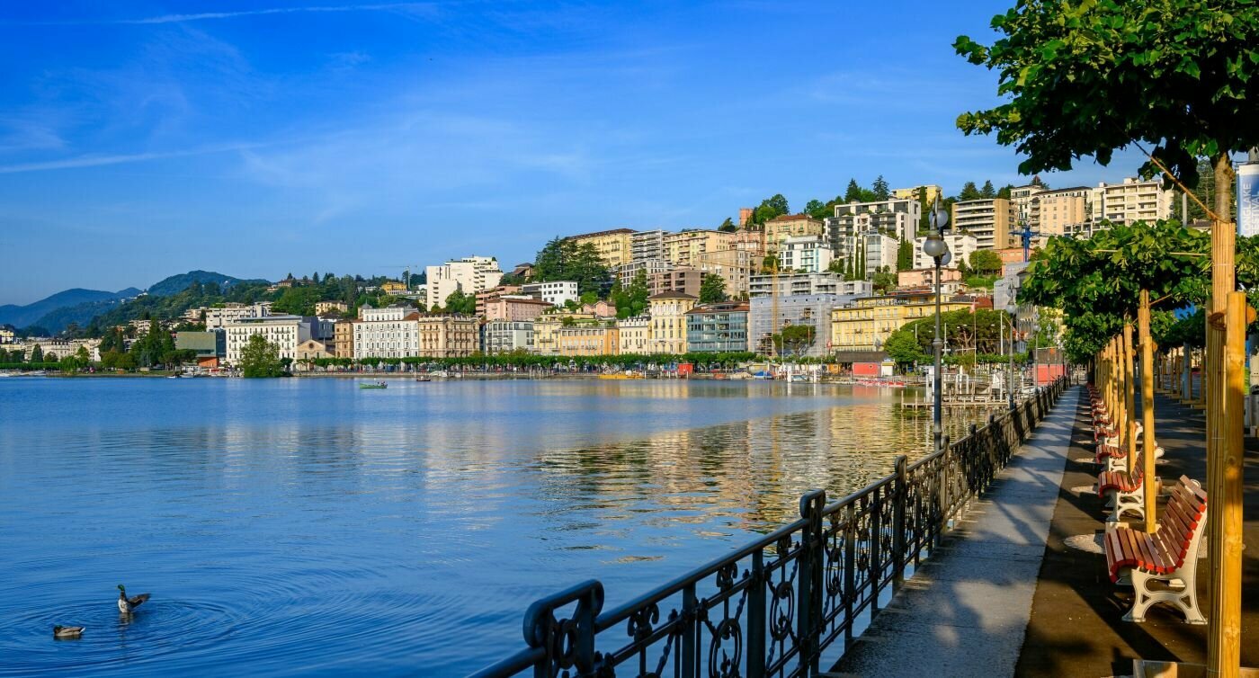 Häuser der Stadt Lugano werden von der Morgensonne erleuchtet und spiegeln sich auf der glatten Seeoberfläche.