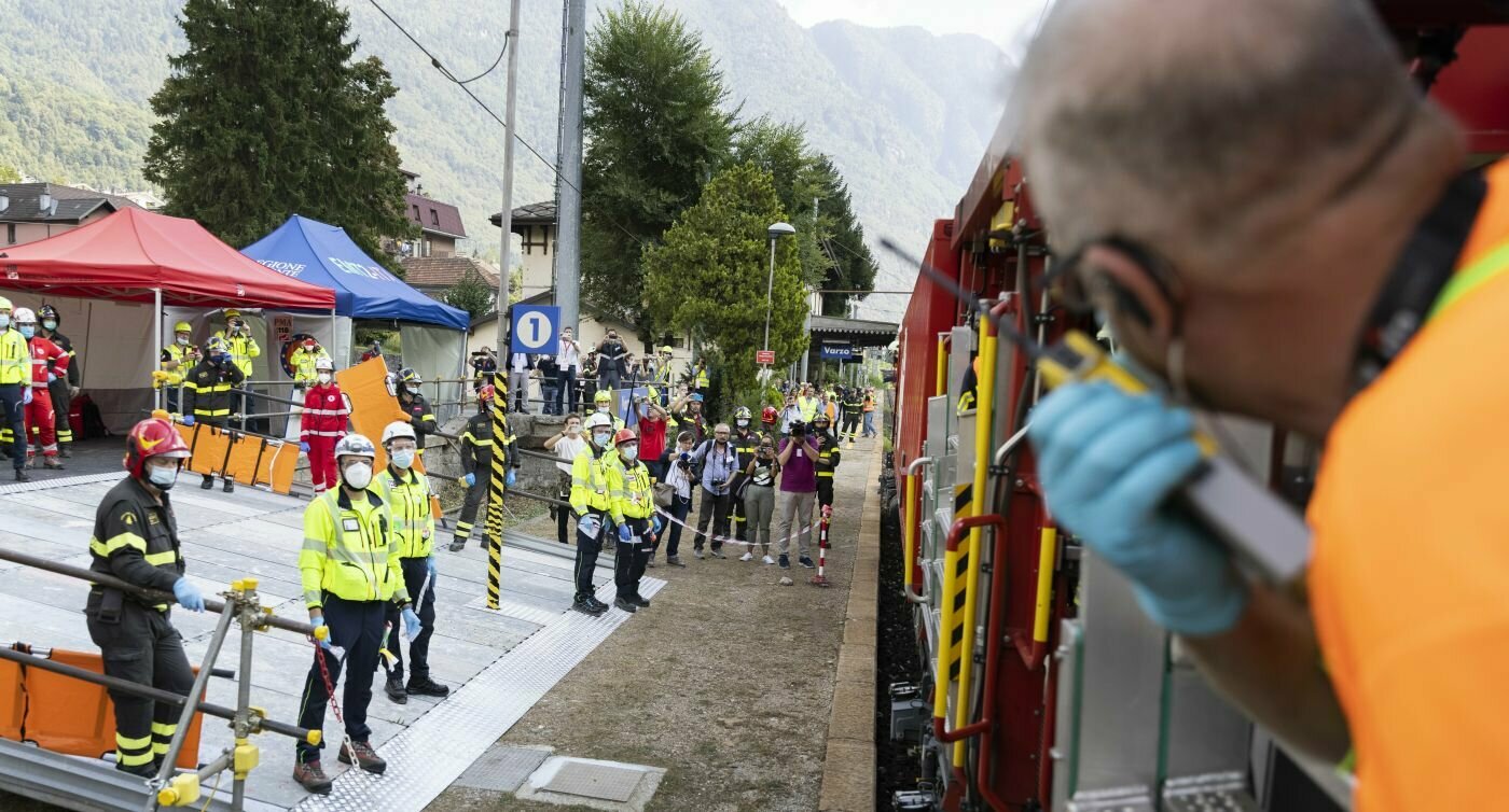 Un uomo si affaccia dal treno e parla in una radio, diversi soccorritori sono in piedi accanto ai binari