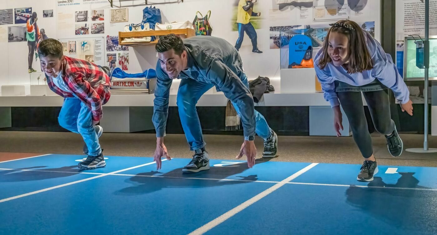Un père, son fils et sa fille prêts à s’élancer sur une piste d’athlétisme d’intérieur bleue au Musée Olympique.