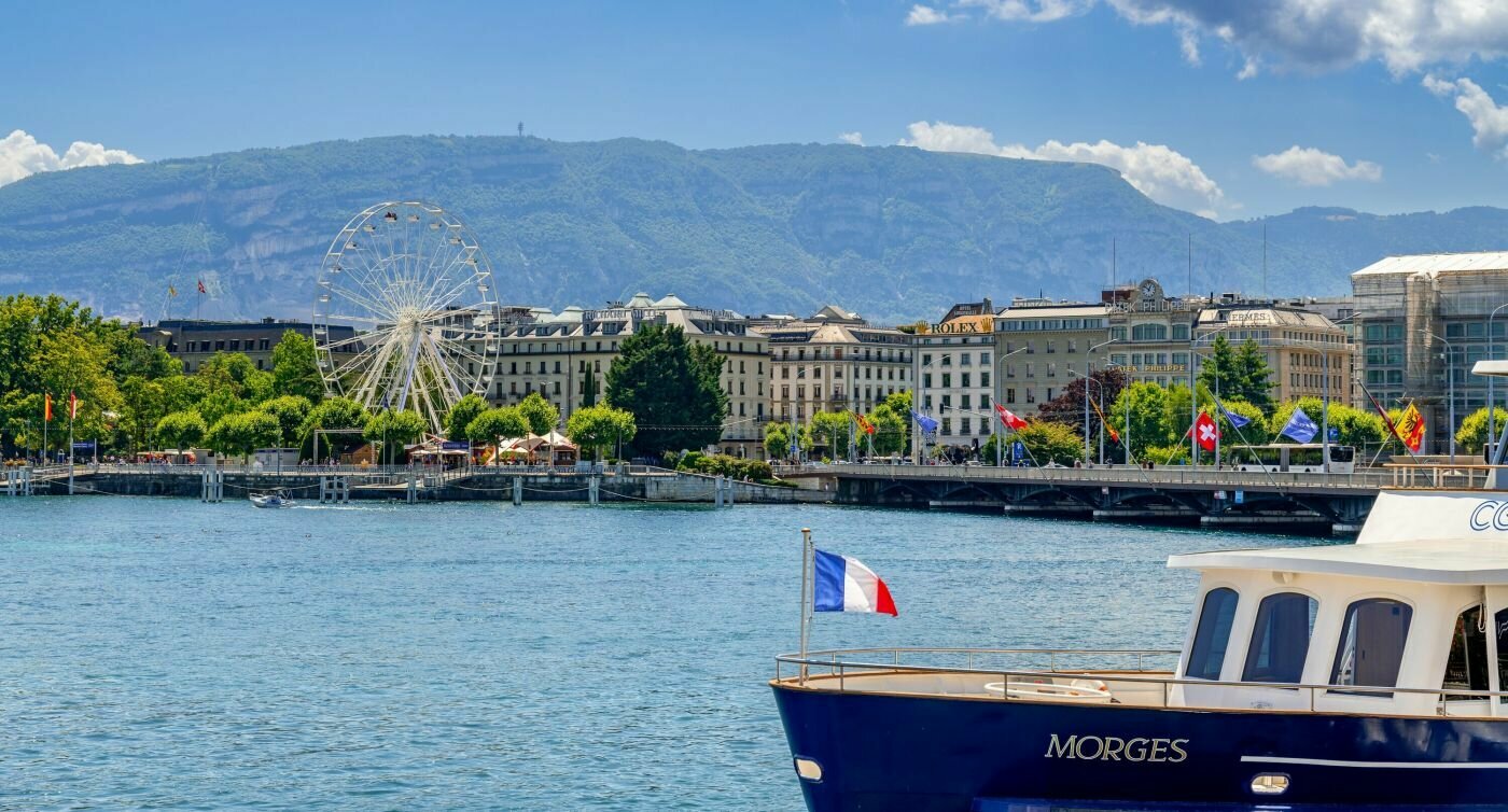 Blick vom Schiff auf die Seepromenade von Genf mit imposanten Gebäuden und einem Riesenrad. 