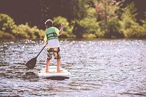 Bambino con giubbotto di salvataggio su uno stand-up paddle sul lago; sullo sfondo un bosco.