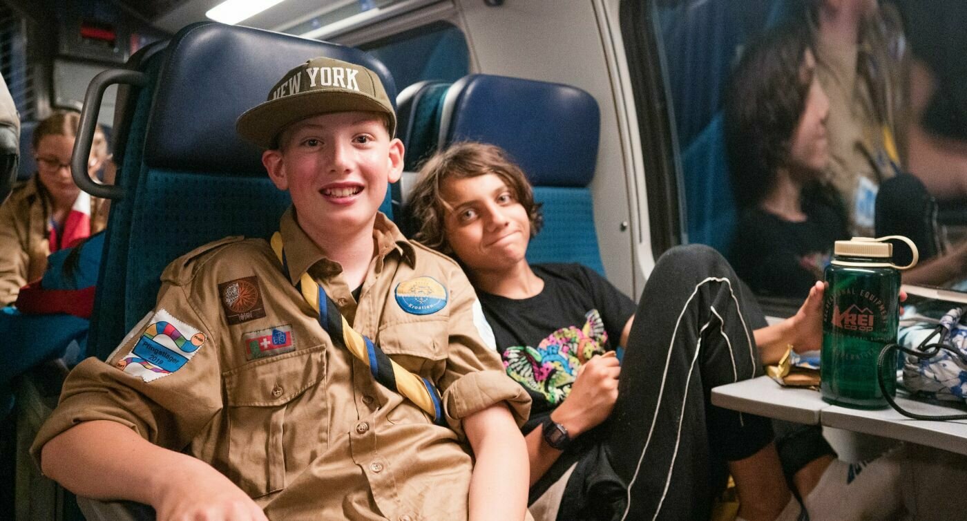 Deux scouts sont assis dans le train et regardent la caméra avec malice.