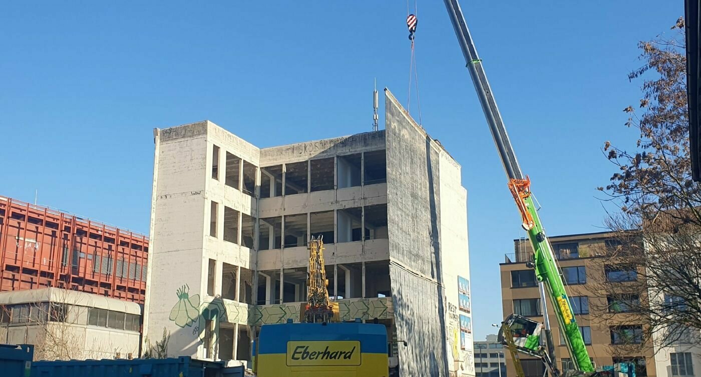 Das Foto zeigt einen Bagger, welcher Abbrucharbeiten an einem mehrstöckigen Gebäude vornimmt, rechts flankiert von einem Pneukran mit Schutzblache. 