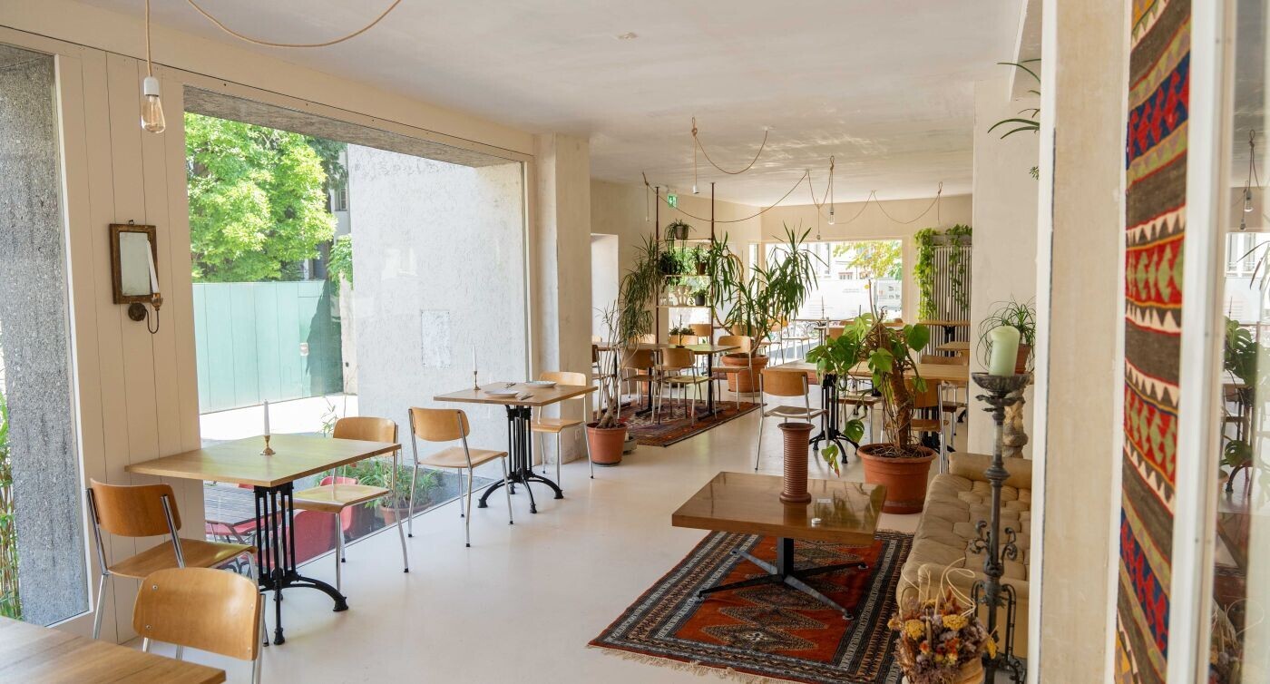 Das Restaurant mit Pflanzen, Tischen Stühlen und einem grossen Fenster.