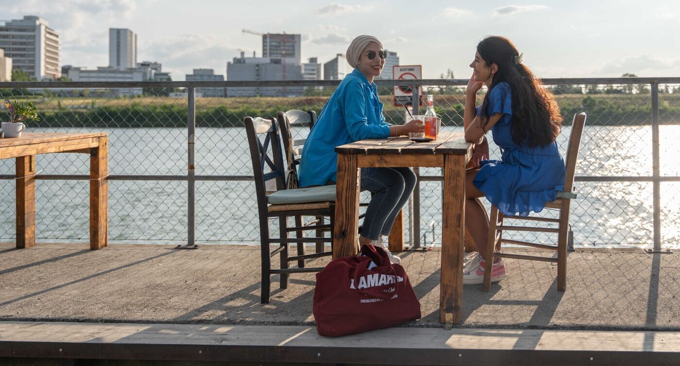 Zwei Frauen sitzen im Restaurant Landestelle am Ufer des Rheins vis a vis voneinander an einem Tisch. 