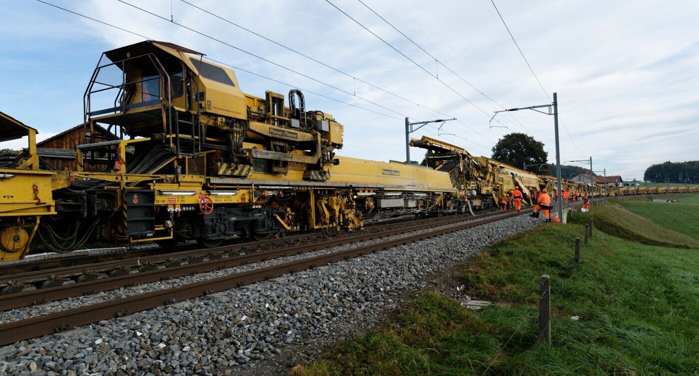 Eine gelbe Gleisbaumaschine und Arbeiter auf offener Strecke. 