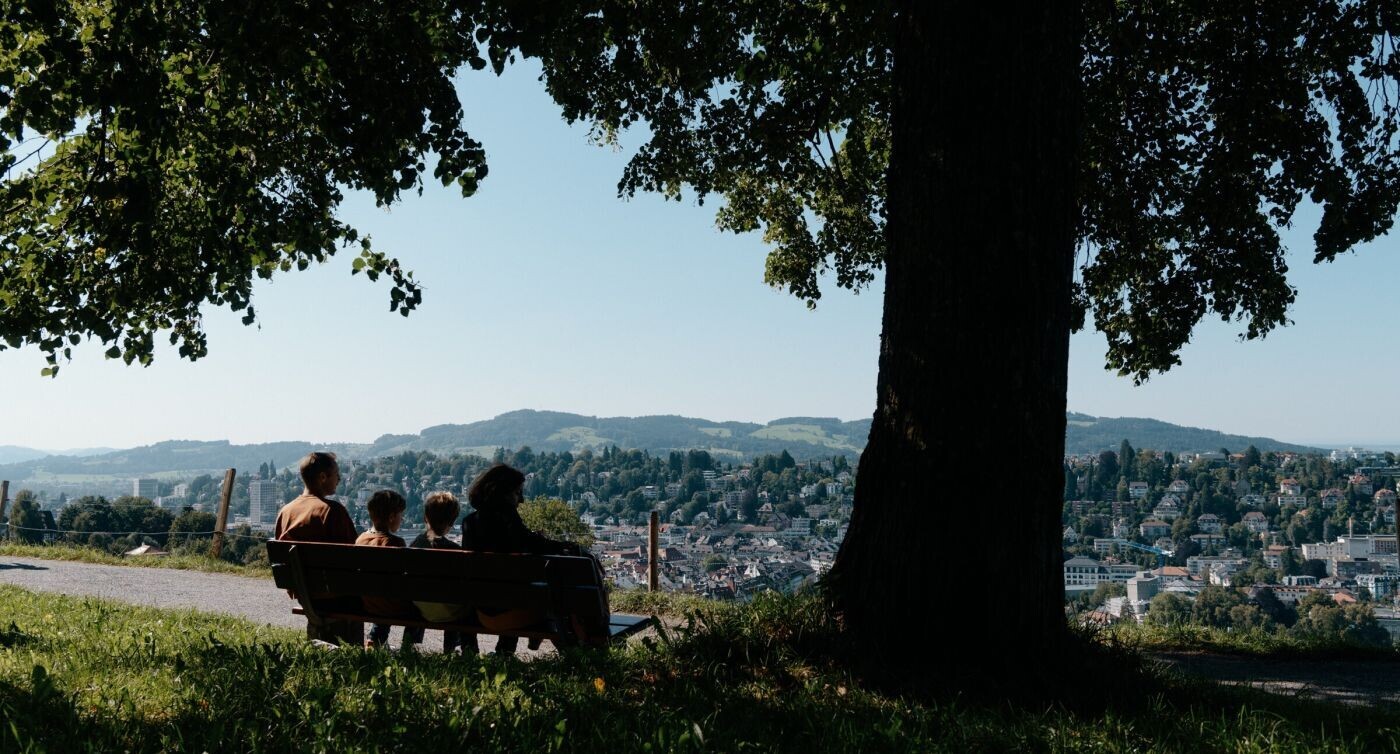Eine Familie sitzt auf einer Bank unter einem Baum und schaut in die Landschaft. 