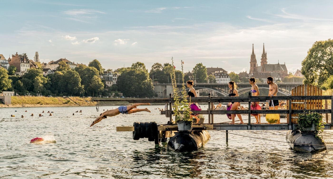 Un homme plongeant d’un ponton dans le Rhin, à sa suite, d’autres personnes attendant leur tour. La vieille ville de Bâle apparaît en arrière-plan. 